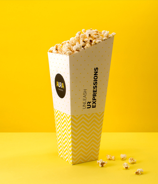 Aura Cinemas popcorn packaging in yellow with the slogan 'Unleash Your Expressions' featuring a modern design and chevron pattern, with a black logo.