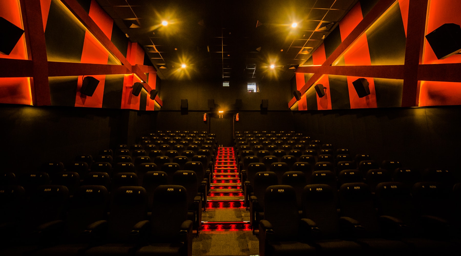 Interior of an Aura Cinemas movie theater auditorium in Bengaluru, with rows of dark seats, red ambient lighting, and illuminated stairs, ready for a film screening.