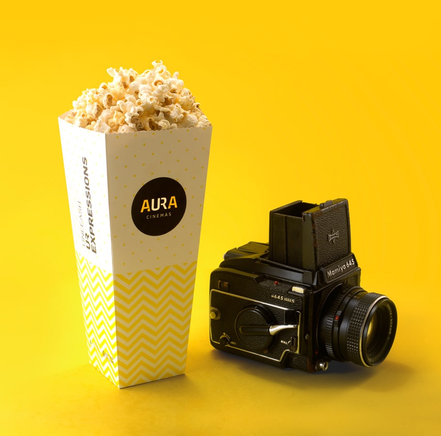 Aura Cinemas branded popcorn in a yellow zigzag-patterned container alongside a vintage camera, capturing the essence of a cinematic experience
