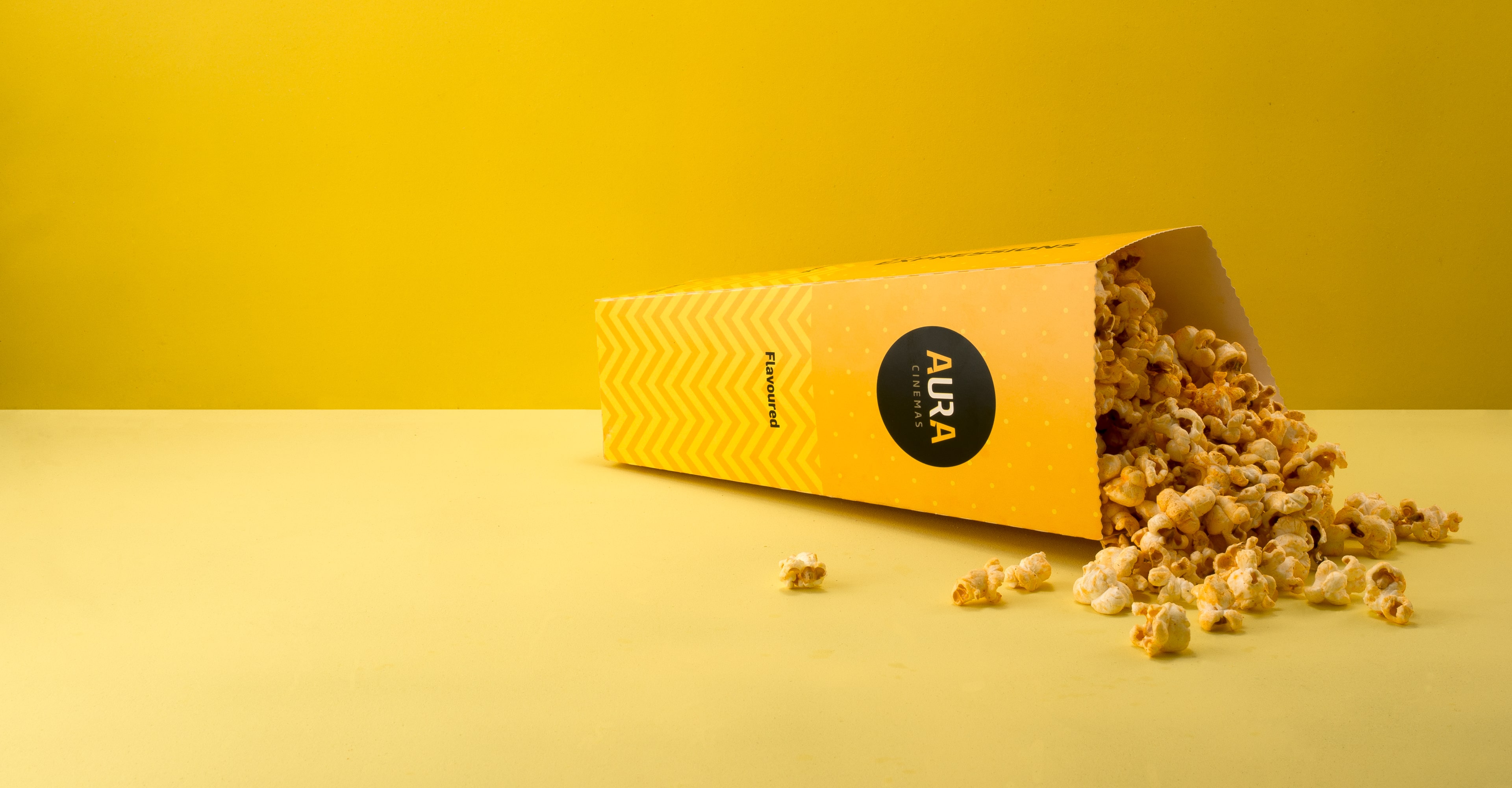 Aura Cinemas flavored popcorn spilling from a yellow zigzag-patterned container, adding a fun twist to the cinema snack experience