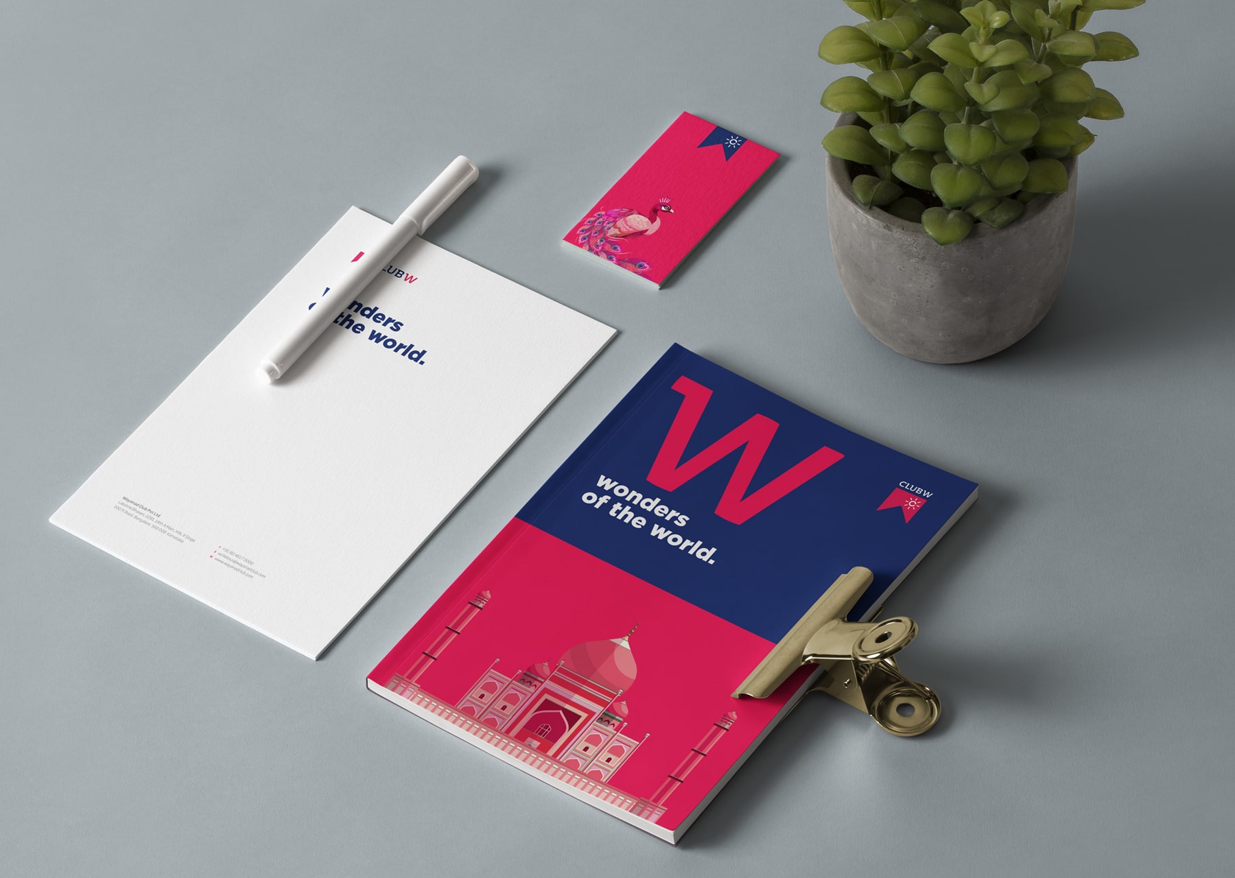 Club W stationery set featuring 'Wonders of the World' notebook with Taj Mahal illustration, matching letterhead, white pen, peacock-themed business card, and potted plant arranged on gray surface