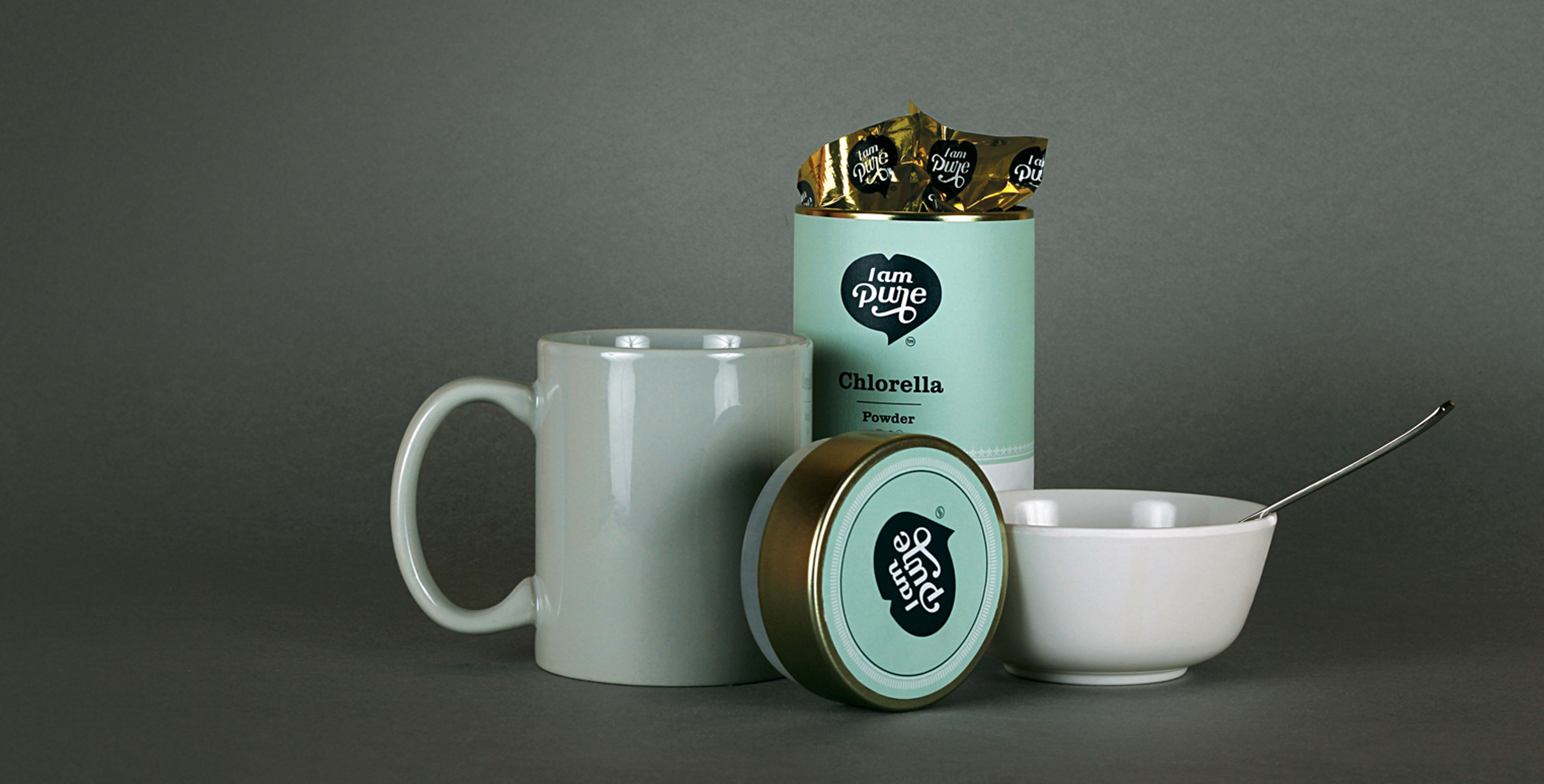 A lifestyle product shot of a I am Pure Chlorella Powder tin, with its lid off and individually packaged supplements visible inside. A light gray mug and a white bowl with a spoon are placed next to the tin on a dark gray background.