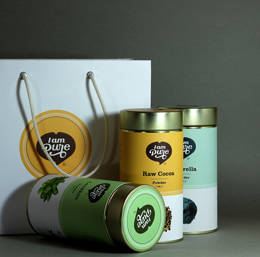 A set of I am Pure brand dietary supplement containers and a white gift bag with the logo. The products shown are Raw Cocoa Powder (yellow), Chlorella Powder (light blue), and Lecithin Powder (green), displayed on a dark gray background.