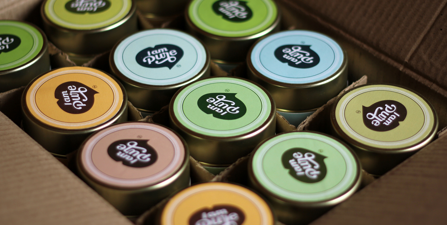 A close-up of I am Pure Super Helse dietary supplement tins in a cardboard box, with lids featuring a variety of pastel colors including yellow, light blue, and green. The brand's logo is prominently displayed on each tin.