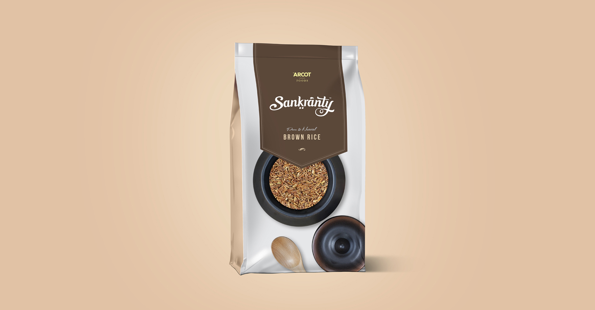 A product packaging pouch for Sankranty Brown Rice. The pouch has a white and brown color scheme with a window showing the grains inside, along with a stylized illustration of a single grain and a pestle and mortar.