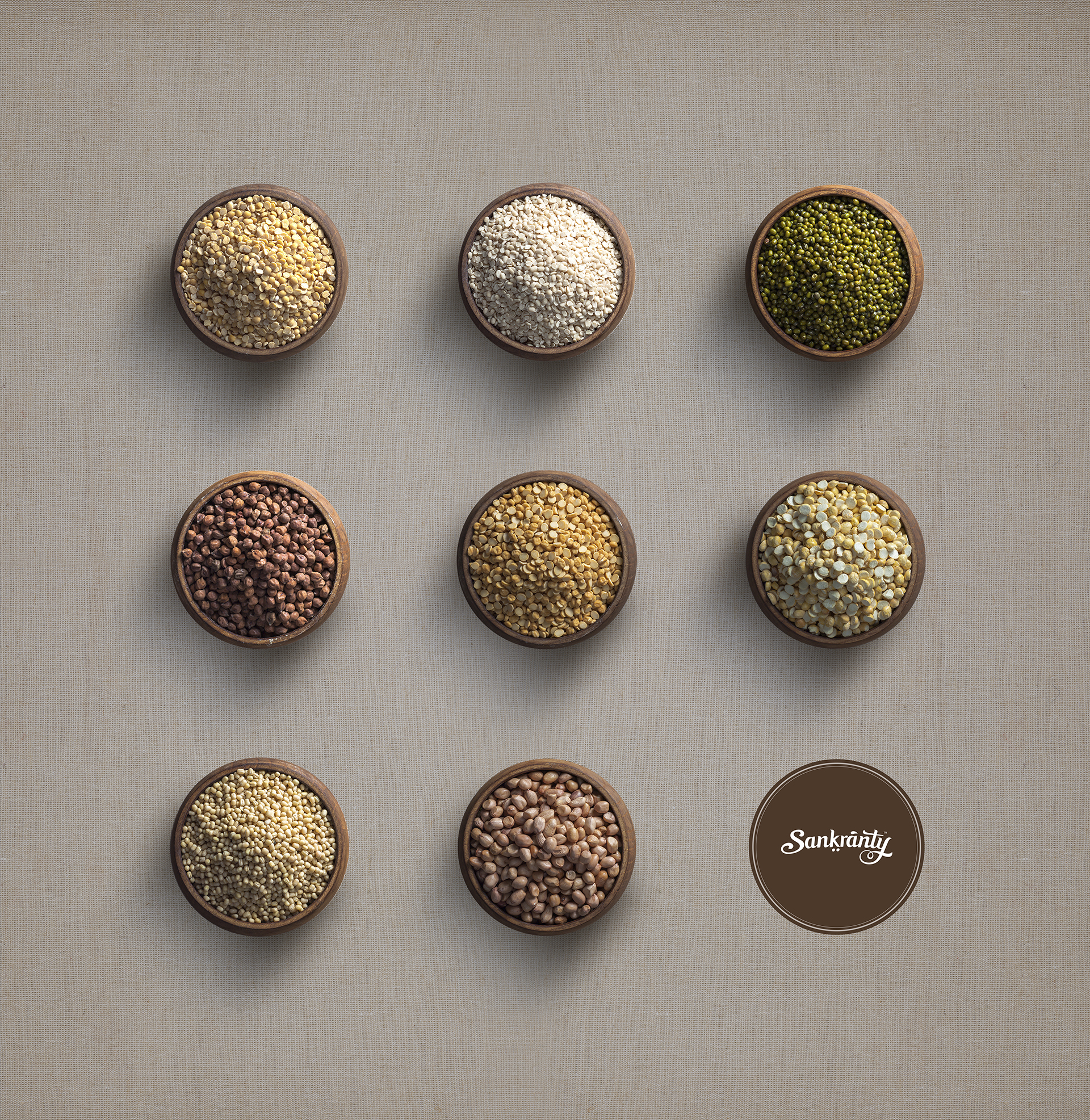 A clean, overhead flat lay of nine wooden bowls filled with different types of pulses and grains, arranged in a grid on a textured light brown background. The Sankranty logo is in the bottom right corner.