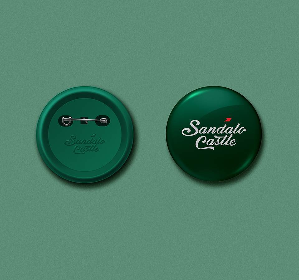 A pair of dark green, circular badges for Sandalo Castle, one showing the front with the logo in white, and the other showing the back with the safety pin and the logo embossed. The badges are on a green background.
