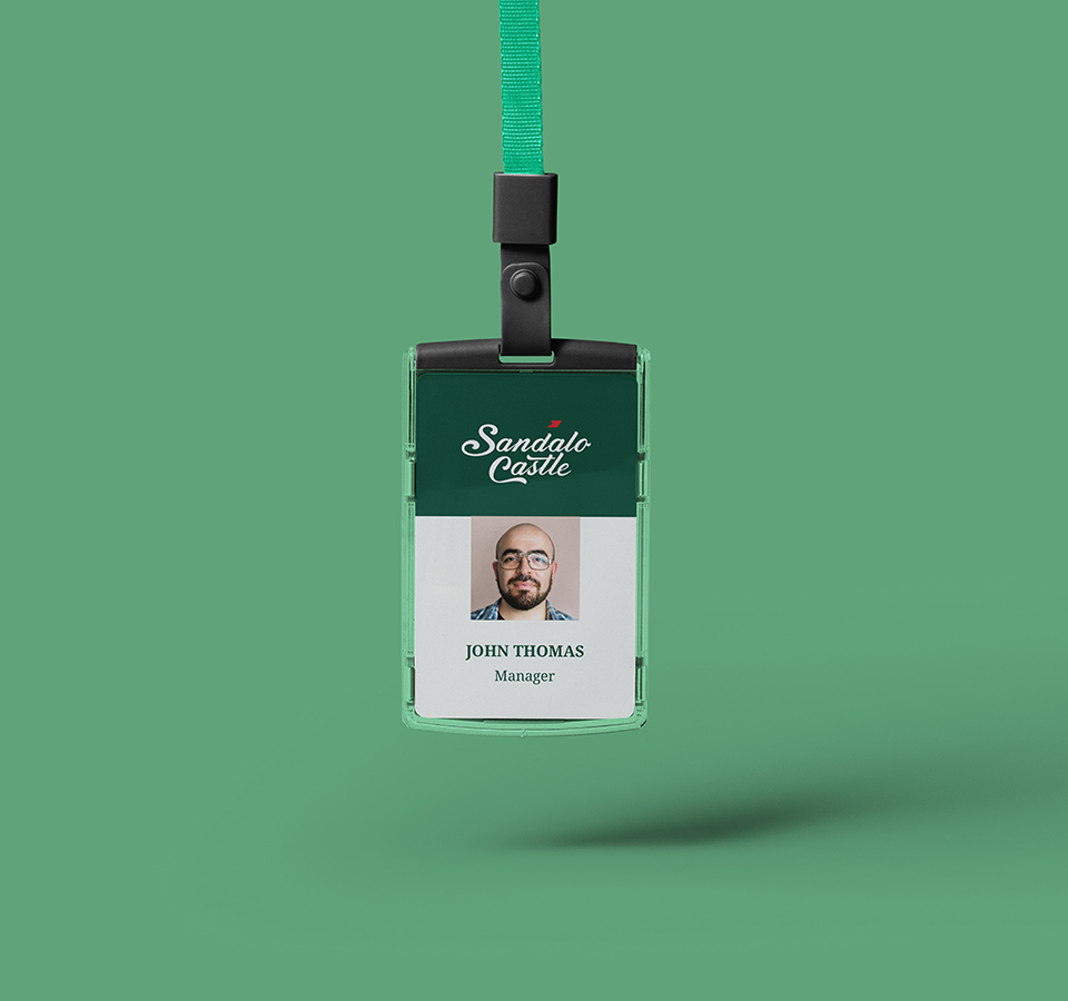 A vertical employee ID card for John Thomas, Manager, from Sandalo Castle. The card features the brand's logo at the top and a photo of a man wearing glasses and a beard, all attached to a green lanyard, floating against a green background.