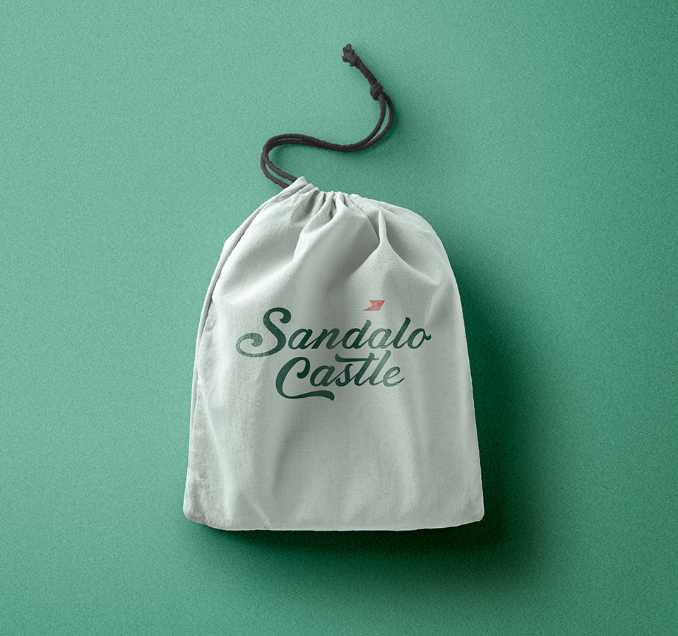 A simple, light-colored fabric drawstring pouch with the Sandalo Castle logo printed on it, lying on a solid teal background. The logo is dark green with a small red flag.