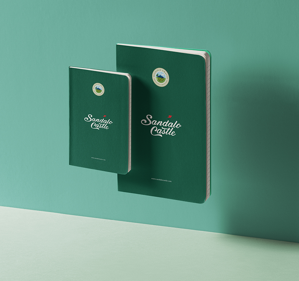 Two hardcover notebooks, one small and one large, for Sandalo Castle. The notebooks are dark green and feature the brand's logo and name, appearing to float against a teal background.