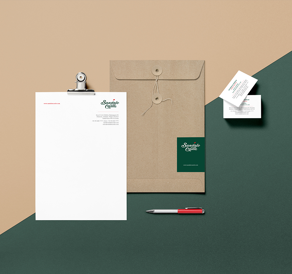 A clean and professional flat lay of branding materials for Sandalo Castle, including a letterhead on a clipboard, a brown string-tie envelope with a branded sticker, business cards, and a red and silver pen, all arranged on a two-toned background.