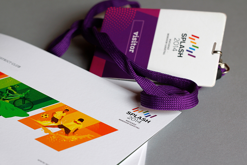 A close-up of marketing materials for Splash 2014, including a visitor's badge with a purple lanyard, and a brochure featuring the SPLASH 2014 Wayanad Monsoon Carnival logo and colorful graphics of people playing sports.