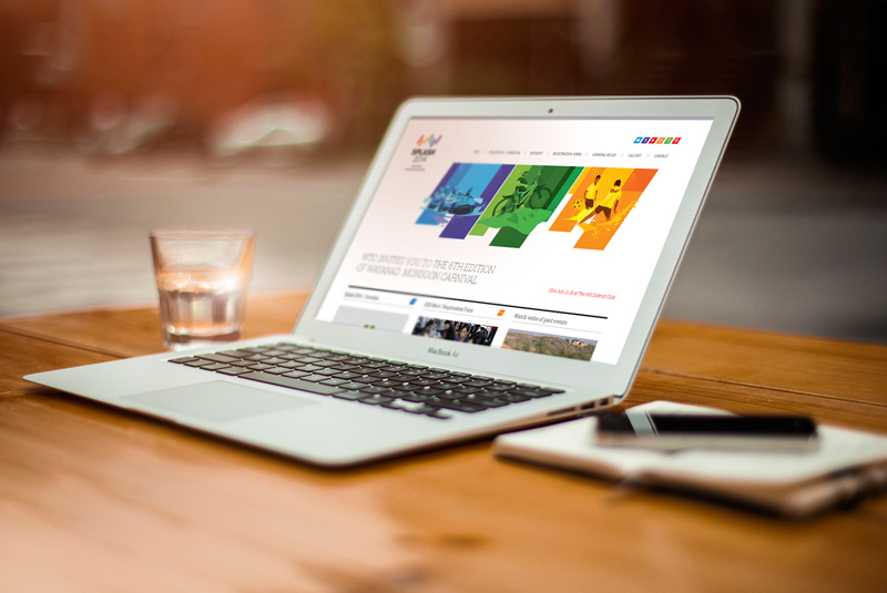 a close-up shot of a MacBook laptop displaying the website for the Splash 2014 Wayanad Monsoon Carnival, showing the event's colorful branding and images, on a wooden table next to a glass of water and a smartphone.
