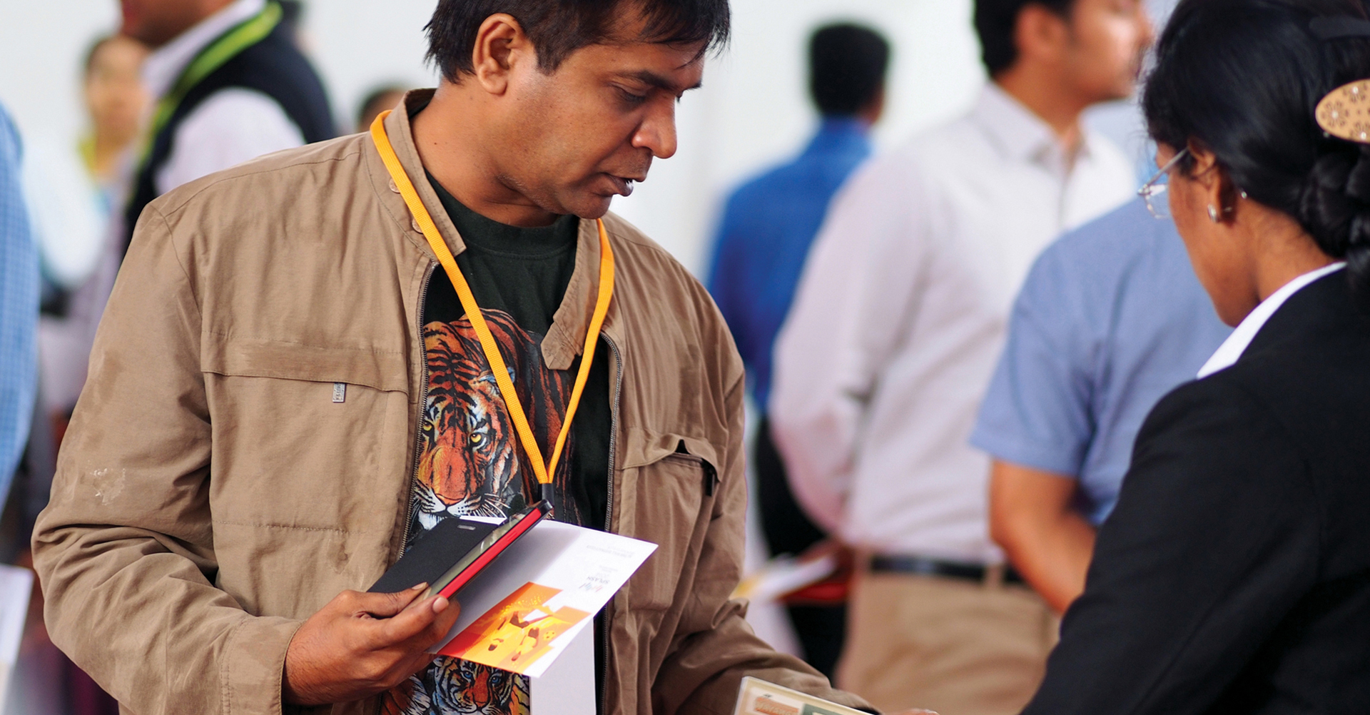 A man wearing a tan jacket and a t-shirt with a tiger graphic is holding brochures and speaking with a woman at a conference or event.