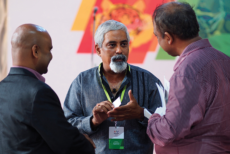 A candid shot of three men in a discussion at the Splash event. The man in the center with a gray beard and hair is wearing a gray shirt and an organiser badge, gesturing with his hands while talking to the two other men.