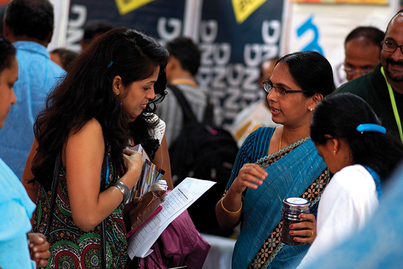 A photo of two women talking at an event booth. One woman, wearing a blue saree and glasses, is holding a small container and speaking to another woman with long dark hair, who is looking down at a form on a clipboard. They appear to be at the Splash event.