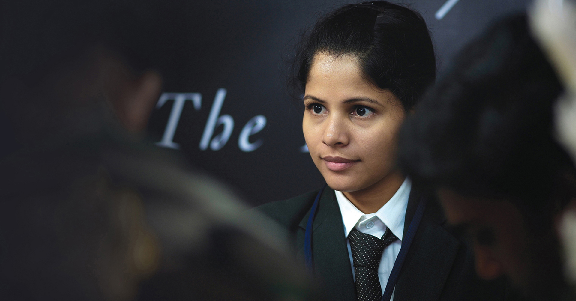 Young woman in uniform looking attentively at a blackboard with 'The' written on it, possibly in a classroom or lecture setting.