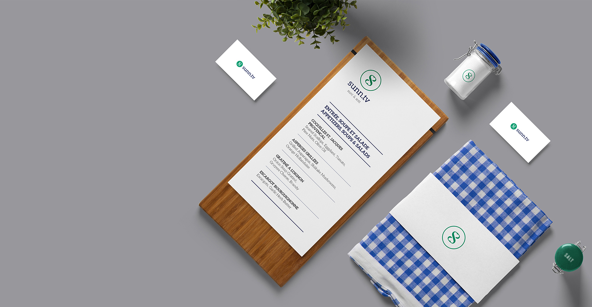 A menu for Sunn.tv is presented on a wooden clipboard. The menu features a clean design with a plant illustration, and the brand's logo is prominently displayed. Beside the clipboard, there is a blue and white checkered napkin with a branded band, a jar of salt, two business cards, and a potted plant, all on a gray background.