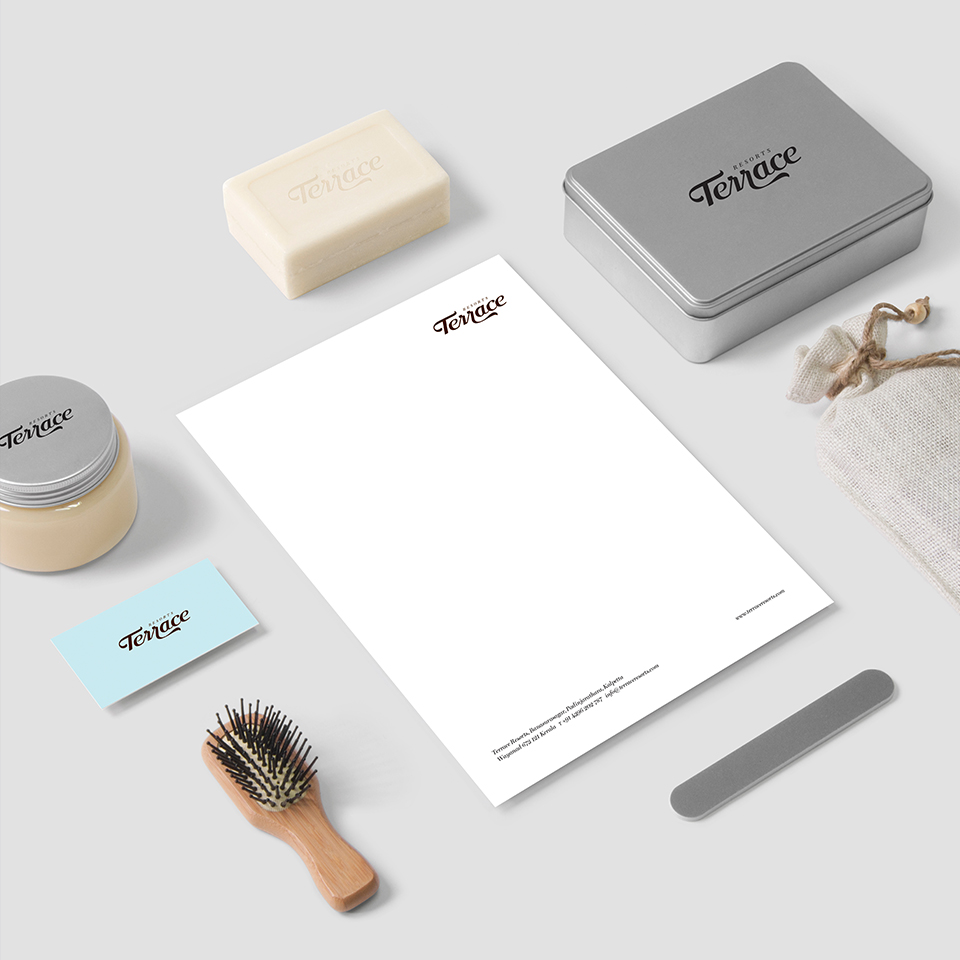 A kitchen flat lay with ingredients and a Terrace Resorts branded menu. The items include a large knife, a wooden cutting board, a bowl of green olives, a tomato, a red chili, a clove of garlic, and a sprig of parsley, all arranged around a light blue menu with a wavy pattern and the resort's logo.