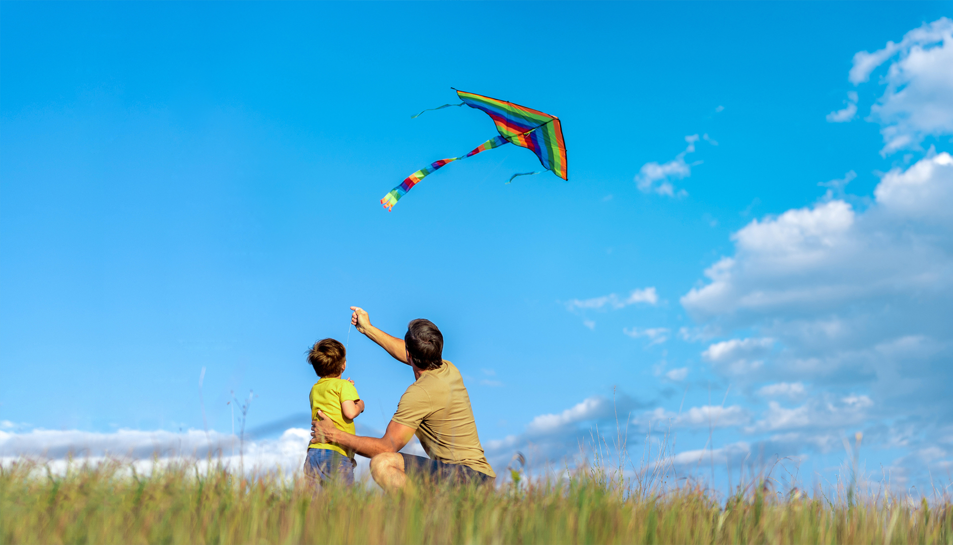 Vacationclub family fun: Father and child flying a colorful rainbow kite in a grassy field under a blue sky.