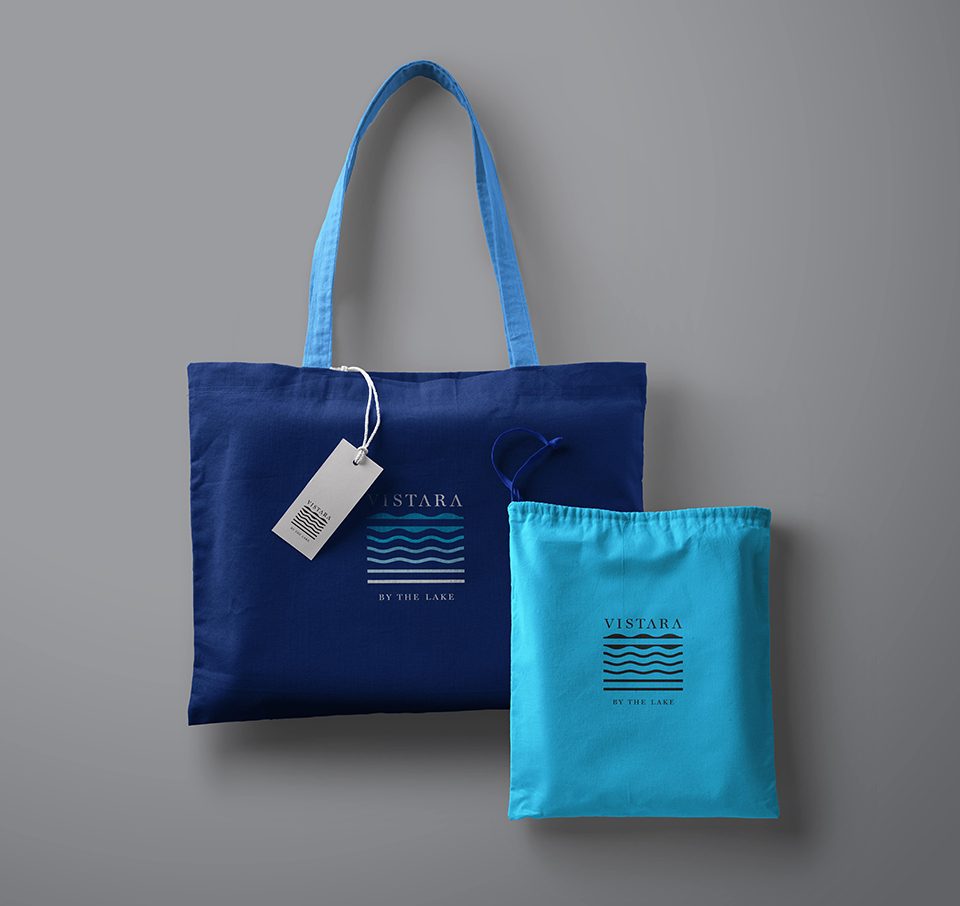Vistara by the Lake branded dark blue tote bag and light blue drawstring pouch with wave-logo design placed against neutral background, showcasing premium resort merchandise