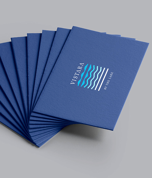 A fan-shaped stack of blue brochures for Vistara, 
                featuring a modern, wavy logo and the tagline 'By The Lake,' representing a resort or housing development.